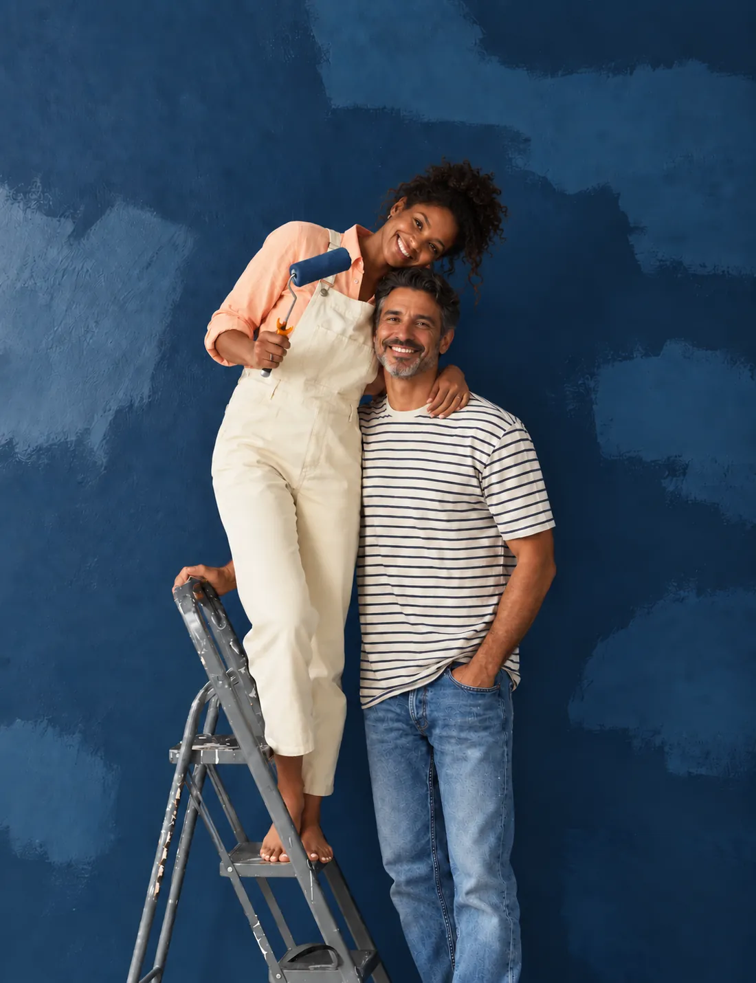 A smiling couple in front of a freshly painted navy blue wall, one standing on a step ladder with a paint roller
